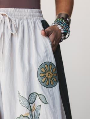 A close-up detail shot of a cream wide-leg pant with intricate floral and medallion embroidery, styled with a stack of beaded bracelets — the kind of piece you reach for when you want your outfit to do the talking.