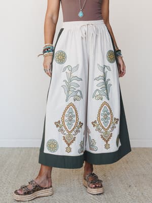 Wide-leg pants in cream with a bold botanical block print and dark olive border panels, styled with a mocha tank and layered jewelry for a well-traveled, intentionally relaxed look.