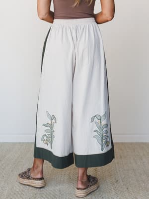 A back-view shot of wide-leg pants in cream with deep olive border trim and botanical embroidery at the hem, styled with a brown tank and leopard print espadrilles — the kind of pants that feel like they came home from a market in another country.