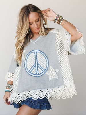 A relaxed oversized top in heathered grey with a bold denim peace sign appliqué and lace star detail, finished with eyelet trim at the hem and sleeves — styled with a ruffle denim mini and stacked bracelets for an effortlessly boho, free-spirited loo