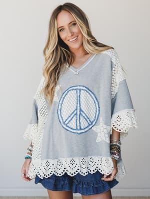 A relaxed oversized top in heather grey with a chenille peace sign appliqué and star patches, finished with eyelet lace trim at the hem and crochet lace sleeves — paired with a denim ruffle mini skirt and layered beaded bracelets for an effortlessly