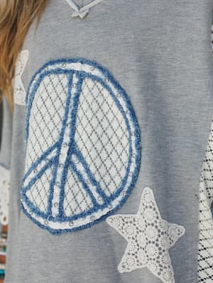 A close-up detail shot of a heathered grey top featuring a large lace peace sign appliqué outlined in blue chenille and rhinestone trim, with crochet star patches scattered across the body — the kind of piece that feels like a vintage find and a new