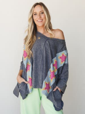 An oversized mineral-washed navy top with bold diagonal chevron panels and hot pink star appliqués styled off-shoulder with neon green pants — effortlessly playful and a little rock-and-roll.