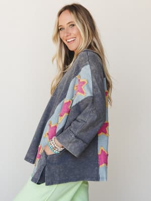 A smiling model shot in three-quarter profile wearing an oversized acid-wash navy top with bold color-block panels and hot pink star appliqués, styled with mint green bottoms and stacked beaded bracelets — the kind of piece that makes a Tuesday feel