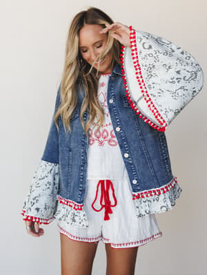 A denim jacket gets a full boho makeover — bell sleeves, animal print ruffle trim, and red pom-pom edging layered over a white embroidered romper with a red tassel drawstring, all shot against a clean studio backdrop with effortless, playful energy.