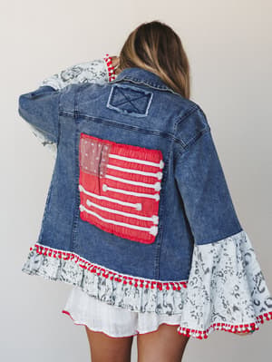 A model shot from behind showing a statement denim jacket with a fabric American flag appliqué on the back, ruffle hem and bell sleeves in a printed fabric, and red pom-pom trim — the kind of jacket that makes people stop you at a Fourth of July part