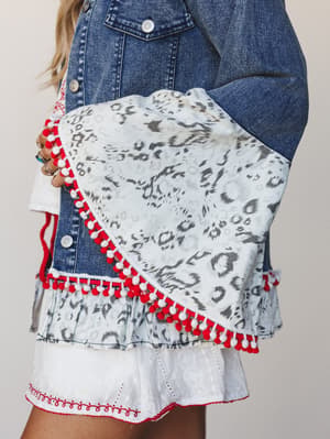 A close-up detail shot of a medium-wash denim jacket layered over a tiered mini skirt, with the focus on playful red and white pom-pom trim along the jacket hem and a leopard-print ruffle detail — the kind of piece that makes a whole outfit feel like