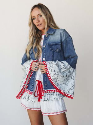 A mixed-media denim jacket with dramatic bell sleeves — the denim body is classic and structured while the sleeves break into a grey animal print ruffle trimmed with red and white pom-poms, worn open over a white eyelet mini dress with red embroidere
