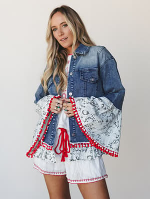A mixed-media denim jacket with dramatic bell sleeves — the denim body is classic and structured while the sleeves break into a grey animal print ruffle trimmed with red and white pom-poms, worn open over a white eyelet mini dress with red embroidere
