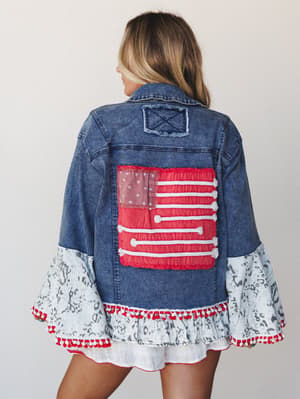 A statement denim jacket shot from the back, featuring a patchwork American flag appliqué, dramatic bell sleeves in a light ikat print, and red pom-pom trim — the kind of piece that makes a whole outfit out of itself.