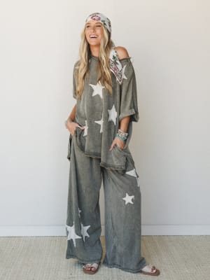 A washed olive matching set with cream star appliqués — oversized top and wide-leg pants worn together with a floral headscarf, sandals, and stacked bracelets, styled with an easy, laughing energy that feels like a really good Saturday.