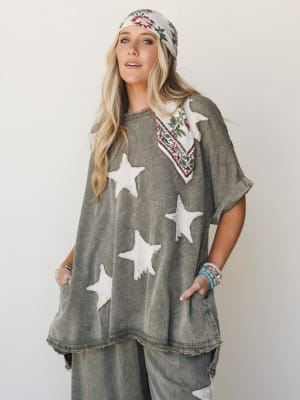 An oversized washed olive poncho top with raw-edge star appliqués is styled with matching wide-leg pants, a floral bandana worn two ways, and stacked turquoise bracelets — the whole look has that perfectly worn-in, I-found-this-at-a-vintage-market en