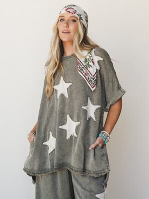 An oversized washed olive poncho top with raw-edge star appliqués is styled with matching wide-leg pants, a floral bandana worn two ways, and stacked turquoise bracelets — the whole look has that perfectly worn-in, I-found-this-at-a-vintage-market en