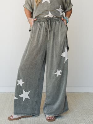 Wide-leg mineral-washed pants with scattered raw-edge star appliqués styled as a matching set, shot against a clean studio wall with a relaxed, lived-in ease.