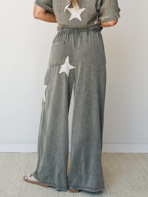 A back-view shot of wide-leg mineral-washed pants in muted olive-grey, featuring chunky star appliqués and an elastic waistband — paired with a matching tee for an effortlessly relaxed co-ord set.