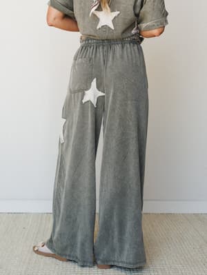 A back-view shot of wide-leg mineral-washed pants in muted olive-grey, featuring chunky star appliqués and an elastic waistband — paired with a matching tee for an effortlessly relaxed co-ord set.