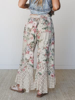 Shot from behind, these wide-leg floral pants steal the scene with a dreamy cream and pink botanical print and a wide crochet lace hem — styled with a denim crop top and sandals for that effortless, garden-party-meets-boho energy.