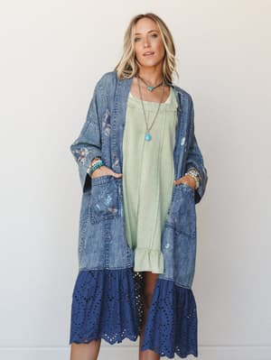 A long denim kimono with floral embroidery and an eyelet ruffle hem is layered over a sage green slip dress — the kind of outfit that looks effortless but clearly had a vision behind it.