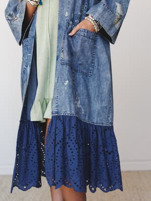 A close-up mid-shot of a layered boho outfit anchored by a long acid-wash denim kimono with subtle floral/bird embroidery, worn open over a soft mint ruffle-hem dress, with a navy eyelet tier peeking out at the hem and stacked beaded bracelets adding