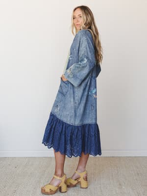 A long denim kimono with scattered floral embroidery and a navy eyelet ruffle hem is styled over a soft green layer, paired with mustard platform clogs — the kind of outfit that makes a Saturday feel intentional.