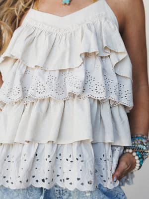 A close-up of a tiered ruffle top in soft cream-taupe, layered with eyelet trim details and paired with distressed denim — the kind of top that makes a casual outfit feel like it had a plan all along.