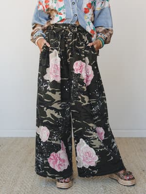 Wide-leg pants in a moody camo-meets-floral mashup — dark olive and black camouflage covered in oversized pink roses and white paint splatter — styled with a denim jacket, floral top layers, stacked bracelets, and platform espadrille sandals for a lo