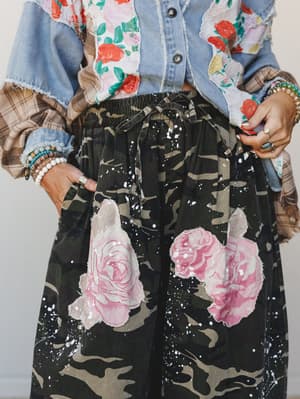 A close-up mid-shot of wide-leg camo pants layered with large pink rose appliqués and white paint splatter, styled under a mixed-media denim and floral patchwork jacket — the kind of outfit that looks like it had opinions before you even put it on.