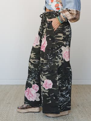 Wide-leg camo pants with oversized pink rose print and white paint splatter details, styled with a cropped denim jacket, plaid sleeves, stacked bracelets, and leopard platform sandals — the kind of outfit that looks effortless but is definitely a who