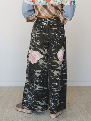 Wide-leg camo pants with hand-painted pink roses and white paint splatter steal the whole shot — styled from behind with a floral crop top, plaid shacket, and espadrille wedges for a look that's equal parts tough and romantic.