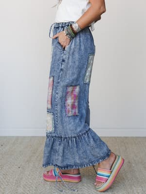 A side-profile shot of a model in acid wash denim maxi pants with colorful patchwork panels and a ruffled hem, styled with a white tee, stacked bracelets, and rainbow platform sandals — free-spirited and full of personality.