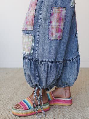 A close-up of wide-leg acid wash denim pants with mixed patchwork panels — floral and plaid in pinks and yellows — ballooning into gathered, tie-hem ankles, styled with rainbow crochet platform sandals on a woven jute rug.