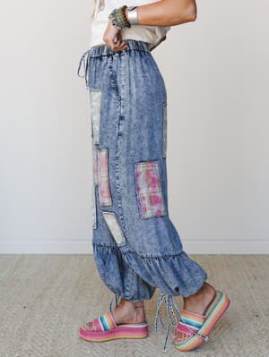A side-profile shot of a model in acid-wash denim wide-leg pants covered in colorful patchwork panels, styled with a cropped cream tee, rainbow platform sandals, and a stack of mixed bracelets — the kind of outfit that looks like it has a whole perso