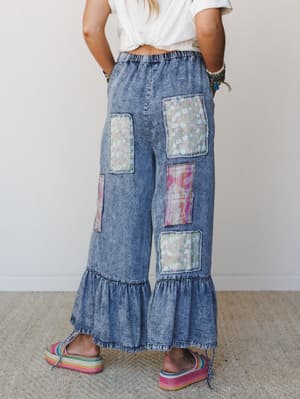 A back-facing model shows off wide-leg acid wash denim pants with mixed floral and abstract patchwork panels, a ruffled hem, and elastic waist — styled with a knotted white tee and rainbow flatform sandals for an effortlessly playful, boho vibe.