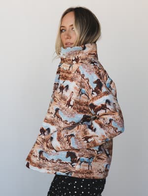 A model glances back over her shoulder in a cropped tapestry puffer jacket covered in a running wild horses scene, paired with black sequin pants — the kind of jacket that announces itself before you do.