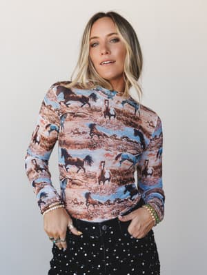 A long-sleeve fitted top covered in an all-over wild horse print — earthy browns, dusty blues, and rust tones — styled with black sequin pants and stacked bracelets for a look that's equal parts Western nostalgia and unexpected night-out energy.