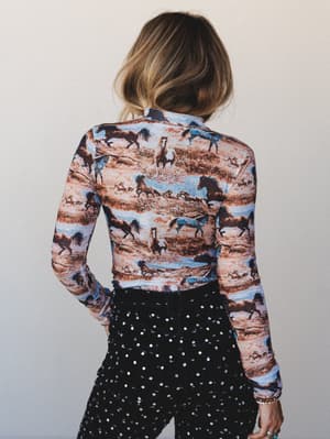 Shot from behind, a model pairs a fitted long-sleeve top covered in an all-over wild horse scene print with studded black pants — the kind of outfit that feels equally at home at a country concert or a girls' night out.