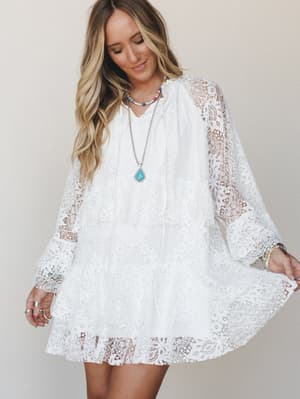 A white lace mini dress with flowy long sleeves and a tiered skirt, styled with turquoise and beaded jewelry for an effortlessly boho feel against a soft neutral studio backdrop.