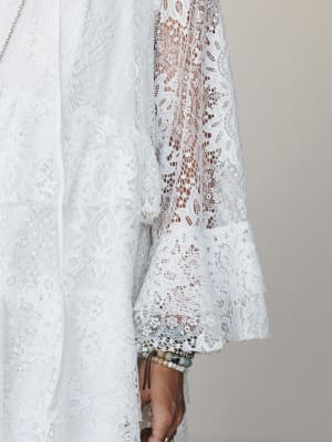 A close-up detail shot of an all-lace dress in off-white, highlighting a sheer floral lace sleeve with a fluted cuff — styled with layered beaded bracelets that feel collected, not coordinated.