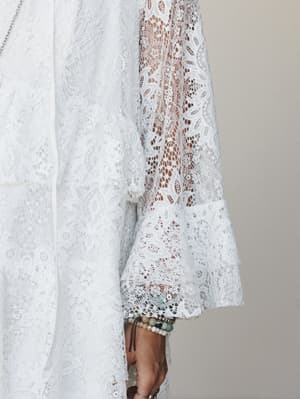 A close-up detail shot of an all-lace dress in off-white, highlighting a sheer floral lace sleeve with a fluted cuff — styled with layered beaded bracelets that feel collected, not coordinated.
