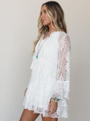 A white lace mini dress with sheer bell sleeves and a tiered scalloped hem, styled with layered turquoise and beaded jewelry against a soft neutral studio backdrop — easy, romantic, and made for warm-weather days.