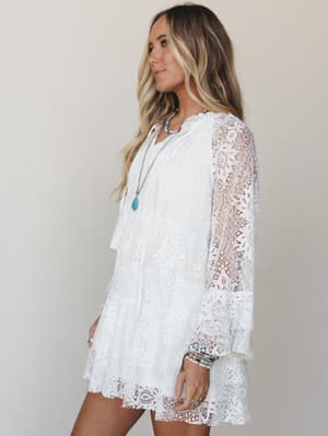 A white lace mini dress with sheer bell sleeves and a tiered scalloped hem, styled with layered turquoise and beaded jewelry against a soft neutral studio backdrop — easy, romantic, and made for warm-weather days.