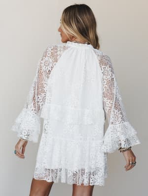 A white lace mini dress shot from the back, all sheer floral lace with tiered ruffle detailing and bell sleeves — the kind of dress that feels equally at home at a boho wedding or a sun-drenched festival.