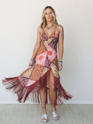 A patchwork bandana-print midi dress with dramatic burgundy fringe hem catches mid-movement against a clean white interior wall — the kind of dress that makes you walk differently the moment you put it on.