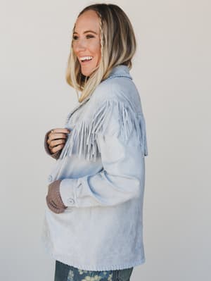 Woman wearing a light blue denim jacket with fringe details on a plain background