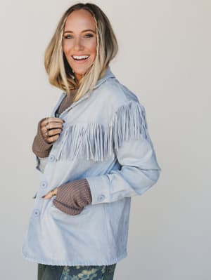 Woman wearing a light blue denim jacket with fringe, smiling against a plain background
