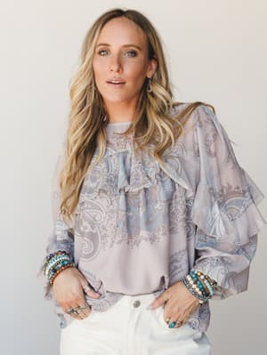 A flowy paisley blouse in dusty mauve and soft blue is styled with white jeans and stacked beaded bracelets — the kind of top that makes a Tuesday feel a little more intentional.