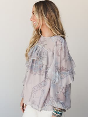 A flowy paisley-print blouse in dusty mauve with tiered ruffle sleeves — the kind of top that looks effortless but clearly has a personality, styled with white pants and a stack of beaded bracelets.