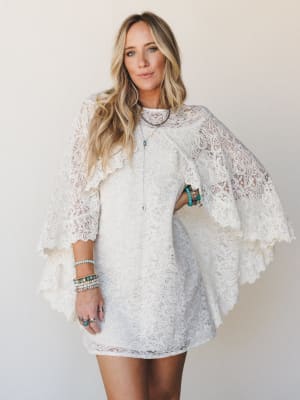 An ivory lace mini dress with dramatic cape sleeves and scalloped trim, styled with layers of turquoise and mixed-stone bracelets and a long pendant necklace — romantic and free-spirited with a hint of vintage bridal energy.