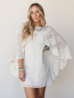 An ivory lace mini dress with dramatic cape sleeves and scalloped trim, styled with layers of turquoise and mixed-stone bracelets and a long pendant necklace — romantic and free-spirited with a hint of vintage bridal energy.