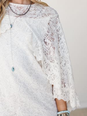 A close-up of an ivory lace dress with wide bell sleeves and scalloped edges, styled with layered turquoise and silver jewelry for a romantic, boho-southwestern feel.